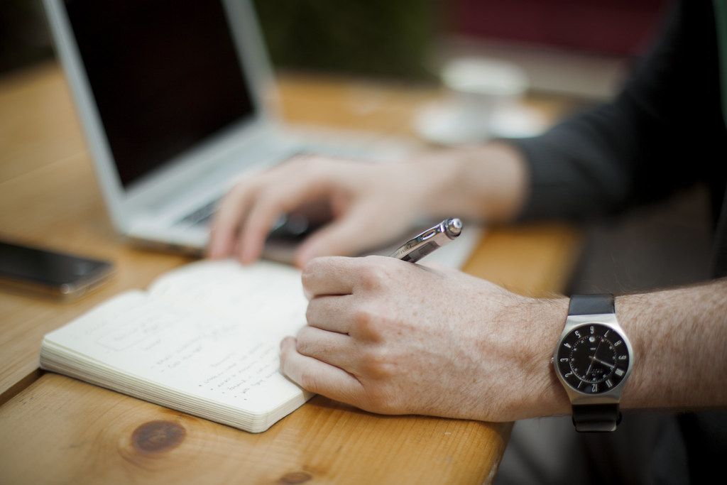 Hand writing notes in a notebook beside an open laptop